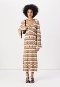 Robe longue ajustée en tissu côtelé, présentant un motif en zigzag dans des teintes de crème, marron et rouille. Décolleté en V avec un accent circulaire et des manches cloche.