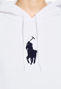 Vit hoodie med en texturerad marinblå broderad polo spelare-logotyp. Designen inkluderar en klassisk dragskohuva och mjukt tyg.
