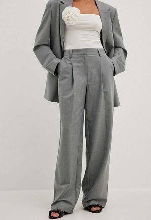 Kvinde iført grå plisserede vidde bukser, matchende blazer, hvid skulderfri top med stor stofblomst og sorte åbne hæle.