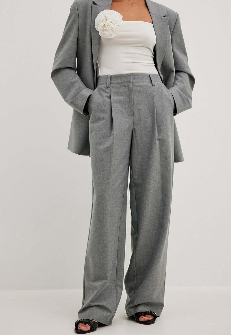 Femme portant un pantalon plissé gris à jambes larges, un blazer assorti, un top blanc bustier avec une grande fleur en tissu, et des talons ouverts noirs.