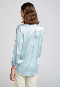 Blouse satin bleu clair avec un motif à poids discrets, manches longues et un détail plissé au dos. Manches avec revers visibles.