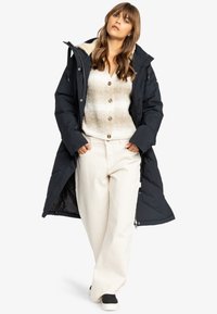 Abrigo de plumas largo negro con capucha de piel sintética, combinado con un suéter de punto crema y pantalones beige claros. Zapatillas completan el conjunto.