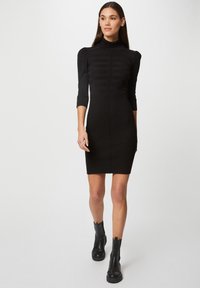 Robe noire côtelée ajustée avec des manches longues, un col haut et un détail de rayures subtiles. Associée à des bottines noires à la cheville.