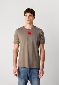 Tricou din bumbac de culoare verde măslin, cu guler rotund și mâneci scurte, având un patch cu logo-ul roșu "HUGO" pe față. Purtați cu blugi albaștri.