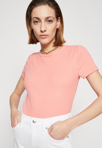 Rosa ribbestrikket topp med korte ermer og rund hals, kombinert med hvite jeans. Stoffet har en jevn tekstur, med figursydde designdetaljer.