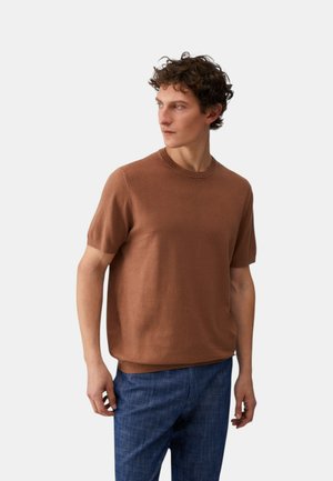 Uomo con capelli ricci che indossa un maglione marrone a maniche corte e pantaloni blu, in piedi e che guarda di lato su uno sfondo semplice.