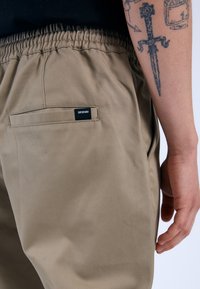 Beige joggers met een elastische tailleband, zijzak en een achterzak met een zwart label. Vlotte stoftextuur en een relaxte pasvorm.