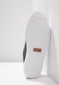 Svart vävd sko med en vit gummisula. Sulan har horisontella ränder och en rektangulär varumärkespatch i brunt.