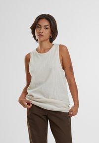 Lys beige uten ermer topp med en teksturert mesh mønster. Rund halslinje og løs passform, sammen med mørkebrun bukser.