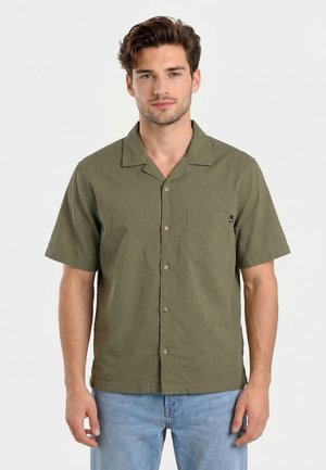 Hombre con camisa verde oliva de manga corta con botones y bolsillo frontal, y vaqueros azul claro, de pie frente a un fondo blanco.