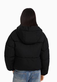 Černá bunda s kapucí typu puffer, s prošívaným vzorem a elastickými rukávy, spárovaná s modrými džínami s vysokým pasem.