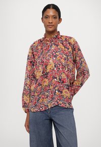 Bluse med blomsterprint i rød, gul og marineblå. Lett materiale, rynket krage og knappestolpe. Kombinert med blå jeans.