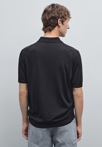 Černé polo tričko s krátkým rukávem a jemnou texturou, s klasickým límcem a hladkým povrchem. Zadní pohled ukazuje přiléhavý střih.