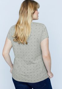 Camiseta corta de manga corta en gris claro con una textura suave, que presenta un patrón de puntos en negro. Con dobladillo ligeramente curvado, combinada con jeans azul oscuro.