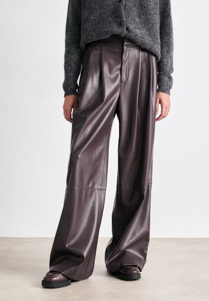 Kvinde iført mørkebrune læderbukser med vide ben, grå fuzzy cropped sweater og mørkebrune chunky sko, stående på et ensfarvet gulv.