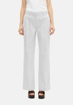 Vrouw in lichtgrijze op maat gemaakte wijde broek met plooien, riemlussen, zijzakken en zwarte plateau sandalen op een witte achtergrond.