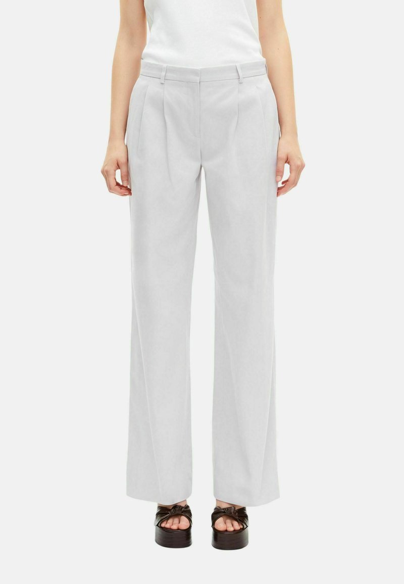 Vrouw in lichtgrijze op maat gemaakte wijde broek met plooien, riemlussen, zijzakken en zwarte plateau sandalen op een witte achtergrond.