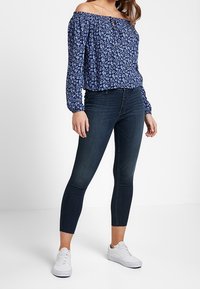 Blue floral off-the-shoulder blouse with gathered sleeves, paired with dark skinny jeans and white sneakers. Smooth fabric and relaxed fit.