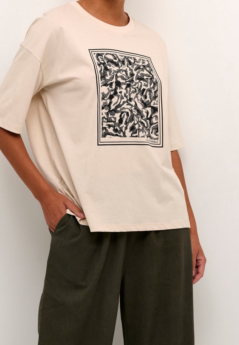 Personne portant un t-shirt ample beige avec un motif abstrait noir et blanc et un pantalon vert olive, une main dans la poche, sur un fond uni.