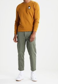 Sudadera de manga larga mostaza con logo de manzana, combinada con pantalones verdes claros. Zapatillas blancas y calcetines completan el conjunto.