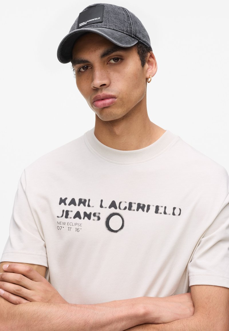 Cappellino grigio con tessuto texturizzato e patch con logo, abbinato a una maglietta a maniche corte color crema con testo grafico e numeri neri.