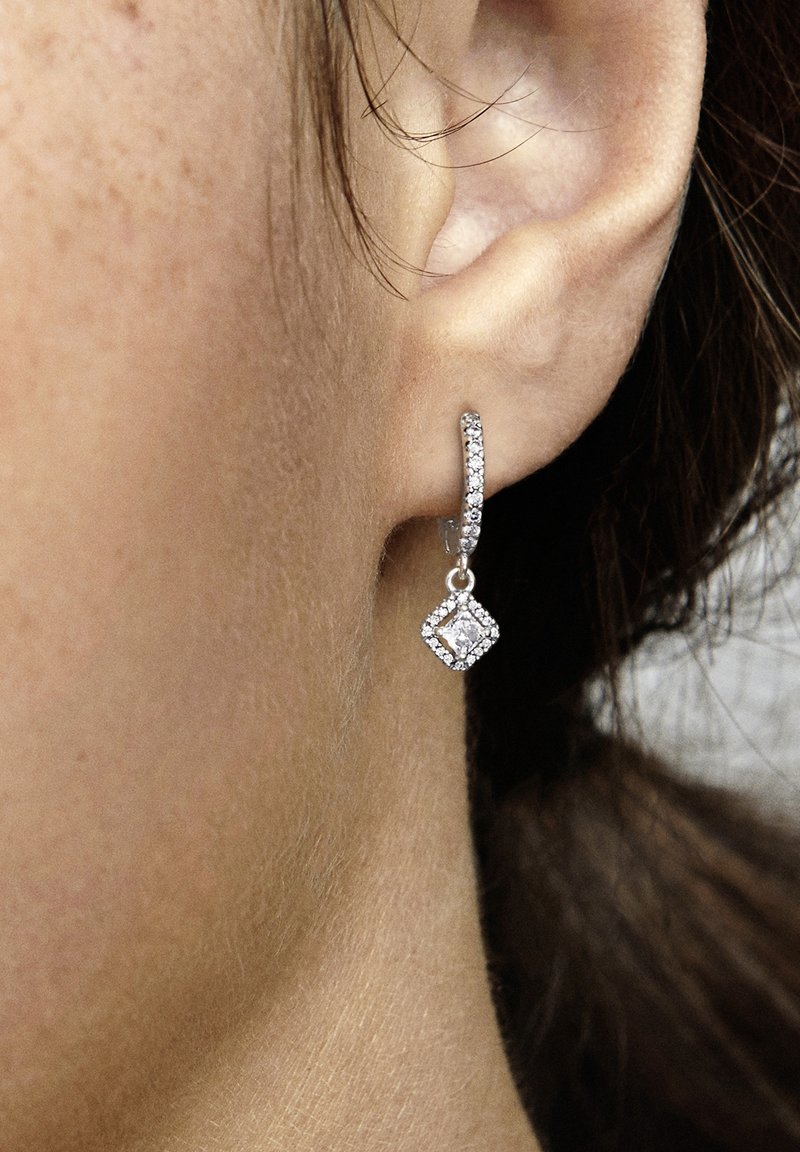 Gros plan sur l’oreille d’une femme portant une boucle d’oreille créole en argent avec de petits diamants et un pendentif carré en diamant suspendu.