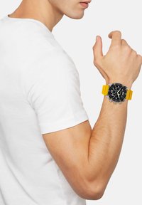 Montre chronographe noire avec bracelet en caoutchouc jaune, matériel en acier inoxydable, et cadran noir comportant plusieurs sous-cadrans, portée sur un bras nu.