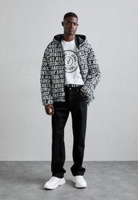 Veste à motifs noir et blanc avec du texte, t-shirt blanc avec logo, jean noir et baskets blanches. Cadre intérieur avec fond neutre.