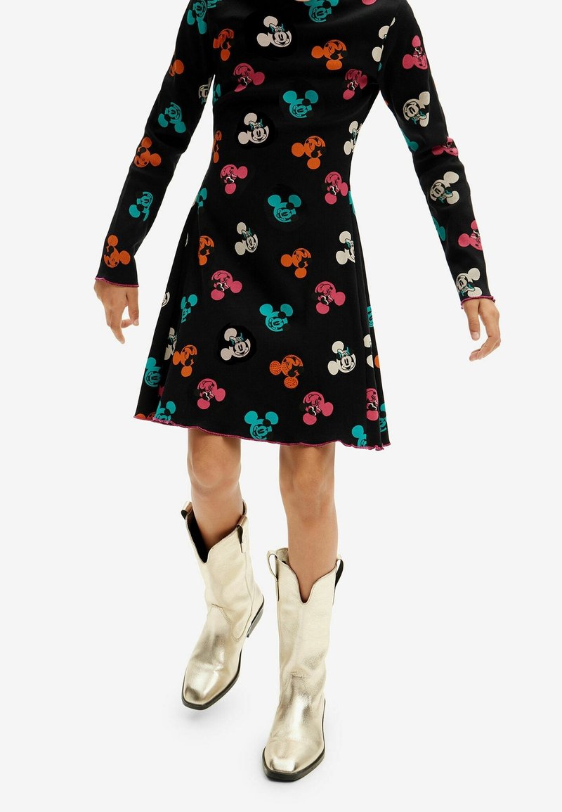 Femme portant une robe noire avec des imprimés colorés Mickey Mouse associée à des bottes montantes argentées métalliques.