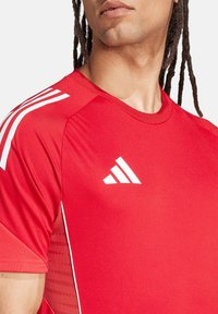 T-shirt de sport rouge avec des rayures blanches sur les épaules et un logo. Texture lisse avec un design ajusté et un motif tonal subtil sur le côté.