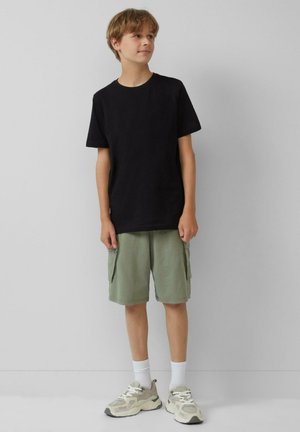 Schwarzes Baumwoll-T-Shirt, grüne Cargo-Shorts mit Seitentaschen, weiße Socken und graue Sneaker mit strukturiertem Design. Schlichte, lässige Kleidung.
