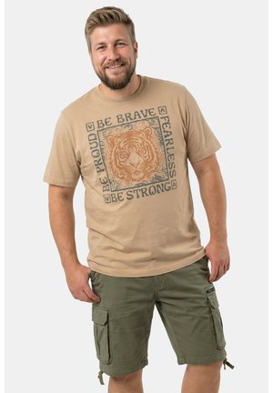 Lächender Mann in einem beigen T-Shirt mit Tiger-Print und motivierenden Worten, kombiniert mit olivgrünen Cargoshorts vor einem schlichten Hintergrund.