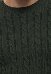 Mörkgrön stickad tröja med en texturerad kabelstickad mönster, ribbad krage och vertikala ränder som skapar en strukturerad design.