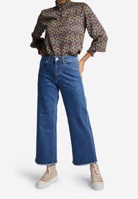 Musterbluse in Braun mit langen Ärmeln und hohem Kragen, kombiniert mit blauen, weit geschnittenen Jeans mit rohem Saum und beigen Schnürsneakern.