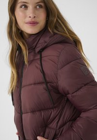 Bordeaux pufferjakke med hætte, der har sorte snore, quiltet tekstur og et logo-patch på venstre skulder. Lynlåslukning foran.