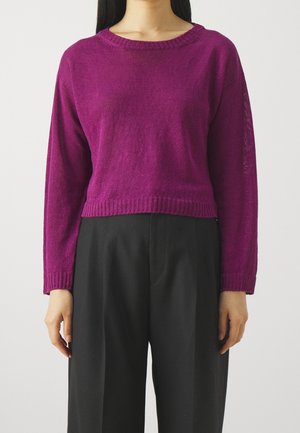 Kvinde iført en langærmet magenta trøje og højttaljede sorte bukser, stående mod en ensartet hvid baggrund.