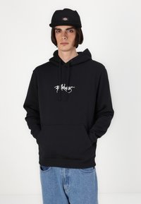 Crna dukserica s kapuljačom i bijelim grafičkim tekstom, prednji džep i vezica. Uparena sa svijetloplavim trapericama i crnom kapom.