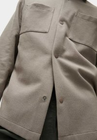 Veste en cuir texturé brun avec deux poches poitrine, boutons-pression et coupe droite. Le tissu semble durable et tissé. La couche intérieure montre une couleur sombre.