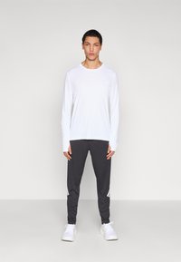 Balta ilgomis rankovėmis sportinė palaidinė, pilkos kelnės su baltomis šoninėmis juostelėmis ir balti sportbačiai, neutraliame fone.