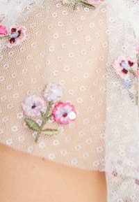 Läpinäkyvä kangas, jossa hennot kukkaompeleet vaaleanpunaisina ja laventelinsävyisinä, pieniä valkoisia pilkkuja ja vihreitä lehtiä vaalealla taustalla.