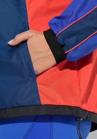 Barevná sportovní bunda obsahující modré a červené panely, texturovanou látku, kapsu s viditelným zipem a černé manžety na zápěstí.