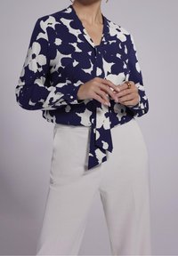 Blouse marine avec un motif abstrait blanc, manches longues et un détail de cravate au cou, assortie à un pantalon blanc. Tissu lisse et coupe ajustée.