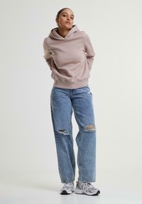 Sweat à capuche en rose clair, en matériau doux. Associé à un jean bleu déchiré avec de grandes déchirures. Baskets grises avec des accents blancs.