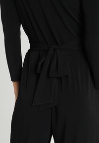 Combinaison noire avec ceinture en tissu nouée à la taille, manches longues et texture douce et lisse, au design épuré.