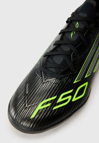 Crampons de football noirs avec un matériau synthétique texturé, des accents vert fluo, des motifs triangulaires et des lacets noirs. Présente le logo "F50" et "SPRINGGRID."