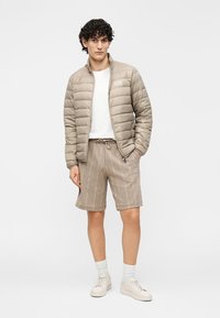 Párnázott bézs dzseki cipzárral az elején, fehér póló és bézs rövidnadrág mintás dizájnnal. Fehér zoknival és világos színű sportcipővel viselve.