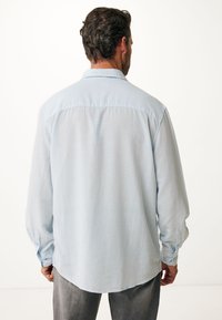 Světle modrá košile s dlouhým rukávem, s límečkem na knoflíky, vyrobená z měkké látky a s zakřiveným lemem. Viditelné jsou zapnuté manžety a hladká textura.