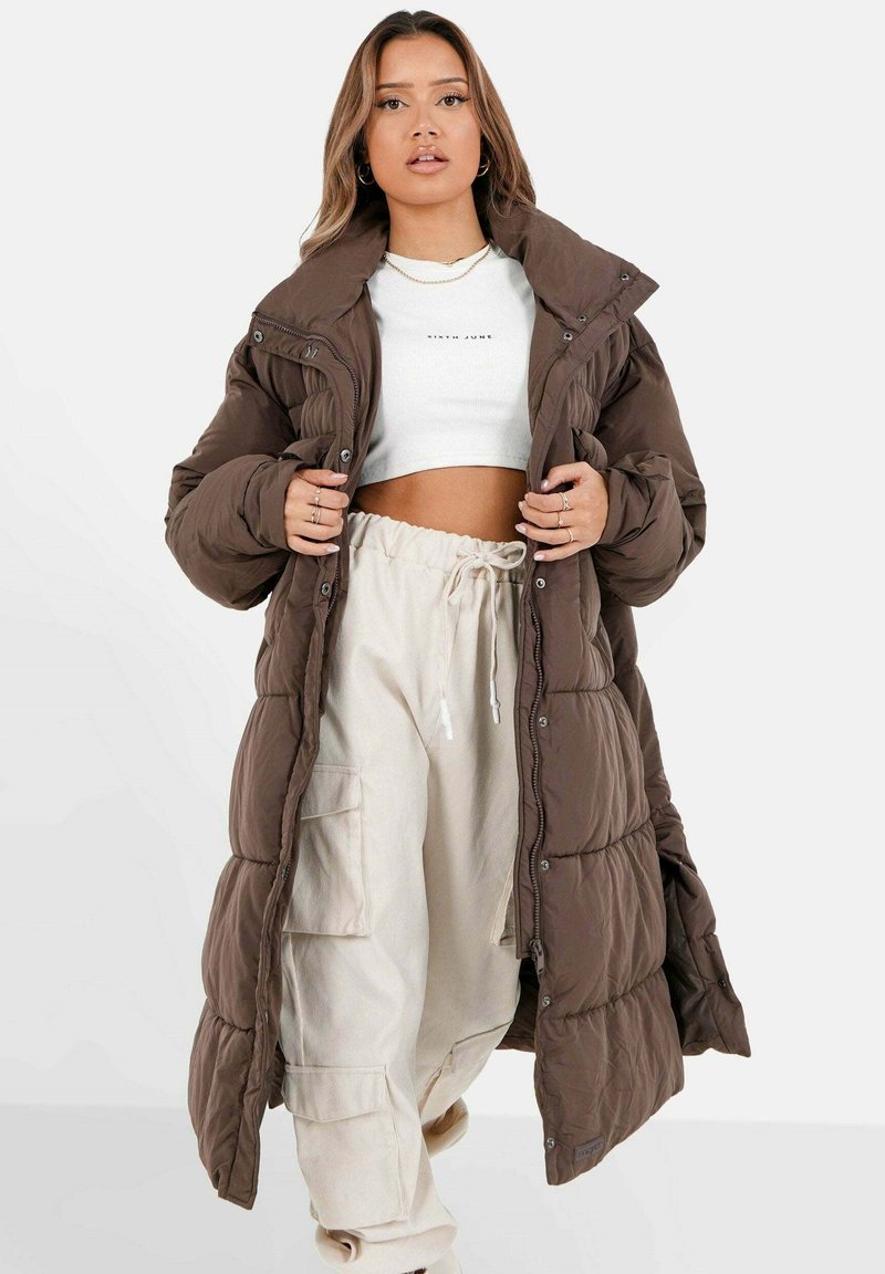 Bruine pufferjas met hoge kraag en drukknoopsluitingen, met een gewatteerd ontwerp. Gecombineerd met een witte cropped top en beige broeken.