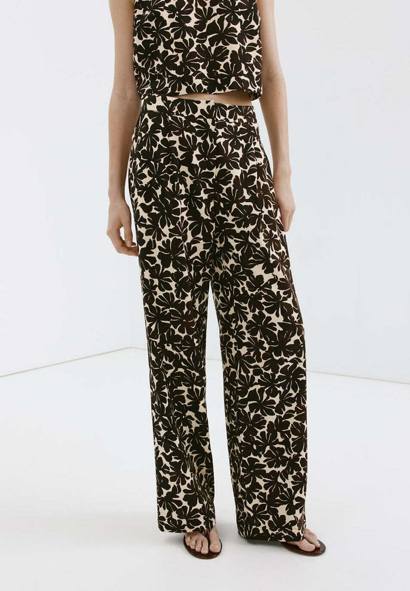 Femme portant un pantalon large à motifs floraux noir et beige assorti et un crop-top sans manches avec des tongs sur fond blanc.