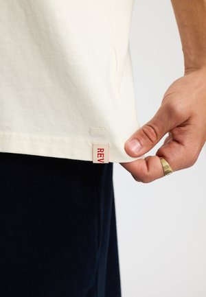 Lichtbeige katoenen t-shirt met een rechte zoom. Een klein rood label dat aan de zijkant is gestikt toont het woord "REV." De structuur lijkt glad.